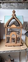 Front view of wooden birdcage showing arched door, wire bars, blue roof panels, finial