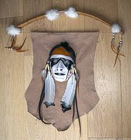 Full view of wall hanging showing bow frame, rawhide panel, mask, feathers