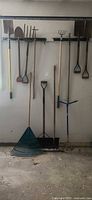 Overall view of ten long-handled garden tools hanging and leaning against wall