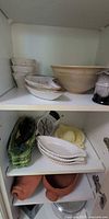 Cabinet shelves showing bowls, corn cob plates, stoneware bowl, salt & pepper grinders, basket dish, apple slicer, citrus juicer, scalloped plates, terracotta bowl, metal pan
