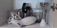 All three appliances arranged in cabinet: white Oster hand blender with cord, stainless shaft, coffee grinder, chopper bowl