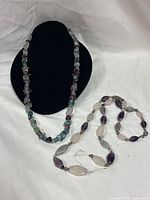 Both amethyst necklaces displayed, chip strand on bust, oval bead strand laid flat