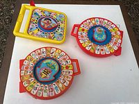 Overview of three See 'n Say toys: two red circular "The Bee Says" units and one yellow Sesame Street unit