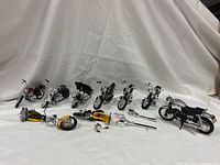 Group of seven complete motorcycles standing with one broken motorcycle and parts in front