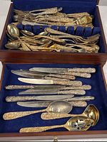 Overview of flatware in wood chest showing upper compartment with assorted gold-tone pieces and lower drawer with knives and serving spoons