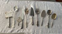 Ten assorted silverplate serving utensils laid out on cloth, overview of shapes and condition