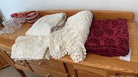 Three folded tablecloths on wooden surface: ivory lace, ivory crochet with fringe, burgundy damask