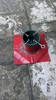 Top view of green and red metal Christmas tree stand with four eye-bolt clamps and red plastic mat