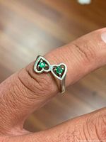 Ring on finger showing two heart-shaped bezels with green emeralds