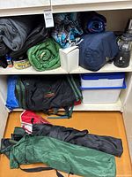 Shelved camping gear including sleeping bags, blankets, lantern, thermos, tote, tarp and chairs in foreground