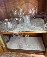 cabinet shelf showing majority of crystal pieces including bowl, pitcher, platter, tumblers, plates, small bowls, ornament and bottom shelf compote and vase