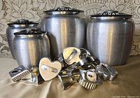 Front view of four Kromex canisters with cookie cutters piled in front