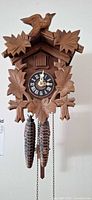 Front view of carved wood cuckoo clock showing dial, bird, leaf carvings and two pinecone weights