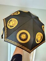 Open black umbrella showing gold Medusa and sun motifs with Greek key border