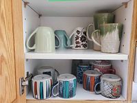 Two shelves in cabinet showing assorted mugs including pastel solids, patterned, and handmade pieces