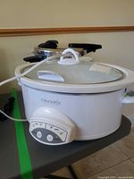 Front view of white Crock-Pot slow cooker showing control panel and glass lid; T-fal pressure cooker partially visible behind