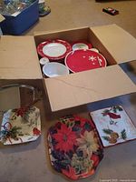 Open box showing assorted red-rimmed plates, bowls, round red snowflake platter, stainless tray and two square holiday platters in foreground