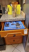 Drawer open with blue tray holding stainless flatware; countertop shows glass utensil holder with wooden spoons and green cutting board