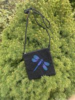 front view of black beaded square bag with iridescent dragonfly and long beaded strap against greenery