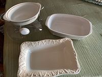 Group view of three white ceramic serving pieces including oval dish in metal stand, plain rectangular dish, and embossed platter