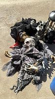 Assorted Halloween decorations on ground: zombie prop, grim reaper, skeletal creature, caged cat, fabrics