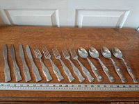 Full set of 13 Coca-Cola pattern flatware pieces laid out on table with ruler for scale