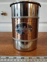 Front view showing two-tone hammered metal utensil holder with UTENSILS plaque and ruler for scale