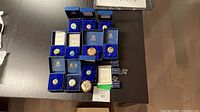 Grouped view of 11 enamel pill boxes in open Halcyon Days boxes with certificates and stands