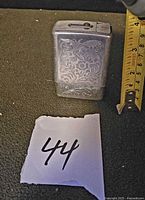 Front view of aluminum cigarette case with floral engraving next to ruler