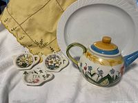 Group shot of all items: teapot, three rests, yellow linen, white platter