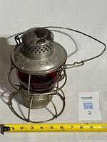 Full view of metal railroad lantern showing red glass globe, wire cage, handle, measuring tape for scale
