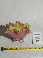 Angled view of entire bowl showing pink exterior, yellow interior, ruffled rim, clear foot, tape for scale