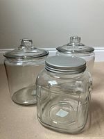 Three clear glass canisters, two round with glass lids, one square with metal lid