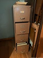 Full view of brown/taupe 4-drawer metal filing cabinet, one drawer partially open showing folders, files on top
