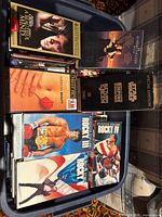 Overview of plastic tote containing multiple VHS tapes; visible titles include Rocky I, III, IV, Star Wars Return of the Jedi Special Edition, A Beautiful Mind, American Beauty plus trilogy slipcase