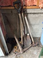 Group of five garden tools leaning against wall: two shovels, pickaxe mattock, hoe, rake