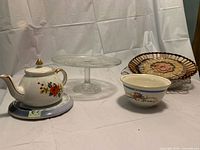 Overall view: teapot on saucer, glass cake stand, serving bowl, wicker basket with doily