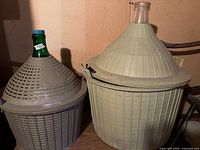 Two demijohns side by side showing size difference, beige plastic baskets, green glass necks