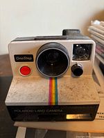 Front view of Polaroid OneStep Land Camera showing lens, shutter button, rainbow stripe, and wear