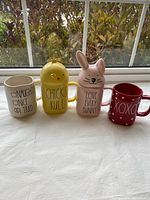 Front view of four Rae Dunn mugs lined up showing text designs and lids