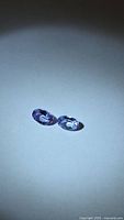 Two oval blue-purple faceted gemstones on neutral background