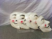 Stack of five identical white Anonymous/Guy Fawkes masks on grey surface with backdrop.
