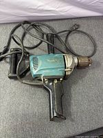 Makita blue corded drill with side handle and power cord displayed on grey surface