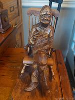 Front view of carved man seated in rocking chair on base