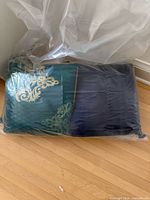 Two of the four pillows visible inside a protective plastic bag, showing green pillow with gold embroidery and blue pillow
