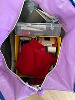 Open purple tote showing yellow box with red fabric, spools of thread, ribbon spool, plastic organizer and other sewing notions