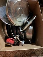 Box filled with stainless-steel cookware, lids, pastry cutter and red plastic accessory