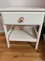 Front view of white wooden nightstand showing drawer, knob, lower shelf