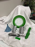 All six kitchen tools grouped: colander, two graters, funnel, zester, peeler, green knife sheath visible