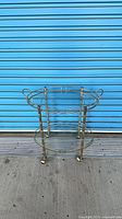 Full view of two-tier brass and glass bar cart standing on pavement, showing handles, shelves, legs and casters
